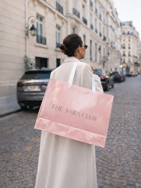 A high-end, soft-focus lifestyle image featuring a branded pink paper shopping bag held by a person in a beige coat on a cobblestone street.